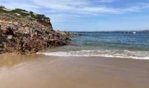 The sandy Bordeira beach with gentle waves rolling in, bordered by rocky terrain and cliffs on the left under a partly cloudy sky.